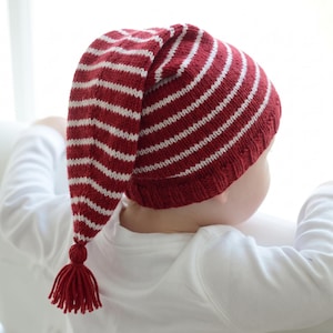May include: A knitted stocking cap featuring horizontal stripes in deep red and off-white. The hat has a long, tapered shape that hangs down, finished with a small red yarn tassel at the pointed end. It includes a ribbed knit brim in solid red.