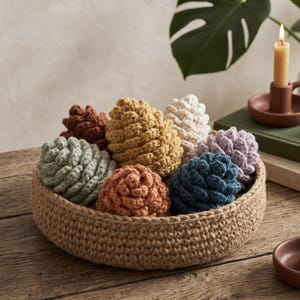 May include: A round, woven jute basket filled with several crocheted pinecone-shaped ornaments in various colors, including mustard yellow, sage green, terracotta, cream, dusty blue, and soft lavender, arranged in a cluster.