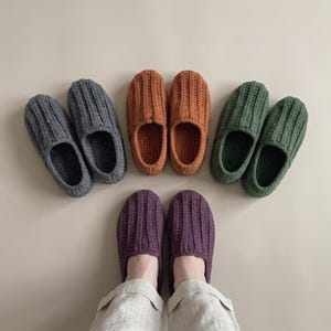 May include: Four pairs of handmade crochet slippers in solid colours including charcoal grey, burnt orange, forest green, and deep purple. Each pair features a ribbed texture throughout the upper and a rounded toe design, arranged in a semi-circle.