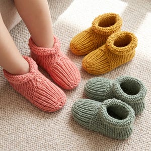 May include: A pair of pink knit slippers worn on feet, accompanied by two additional pairs of knit slippers in mustard yellow and sage green. Each slipper features a thick, ribbed texture, a rounded toe, and a wide, folded-over cuff opening.