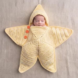 Op de afbeelding: Een lichtgele gehaakte baby-inwikkeldeken in de vorm van een vijfpuntige ster met een capuchon. De inwikkeldeken is voorzien van drie kleine, ronde, feloranje knopen op een van de punten, waarmee de doek rond een slapende baby wordt vastgezet.
