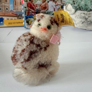 Könnte beinhalten: Plüschkatzenfigur mit weißem und braun gemustertem Fell, grünen Augen und einer rosa Schleife. Die Katze hat ein Etikett mit der Aufschrift "Steiff". Die Figur steht vor einem Reisebuch.