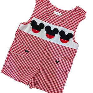May include: A red and white gingham romper featuring a white chest panel with three black and red mouse-ear appliques. The garment includes two front pockets with small black mouse-shaped buttons and red snap closures at the shoulders.