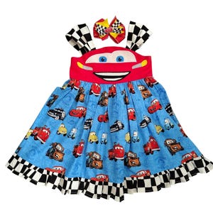 May include: A sleeveless dress featuring a red bodice with a cartoon car face, blue skirt with a repeating pattern of various cartoon cars and the text Radiator Springs, and a black and white chequered hem. Includes a matching hair bow.