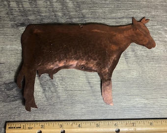 May include: A copper silhouette of a cow, approximately 18 cm long, is displayed on a wooden surface. The cow is facing right and has a slightly raised tail.