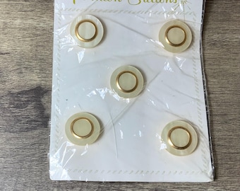 May include: A pack of five white and gold round buttons. The pack is labelled "Unusual Value in Fashion Buttons".