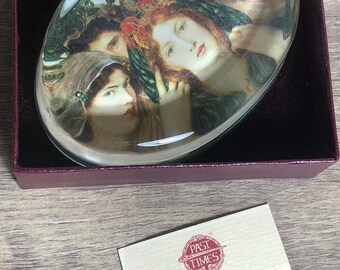 May include: A glass paperweight featuring a painting of four women's faces. The paperweight is oval shaped and has a green border. The paperweight is in a red box with a white card that says "Past Times Oxford England" and includes contact information.