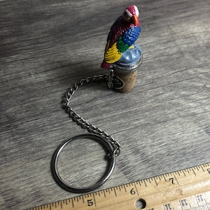May include: A colorful parrot bottle stopper with a metal chain and ring. The parrot is perched on a cork stopper with a silver top.