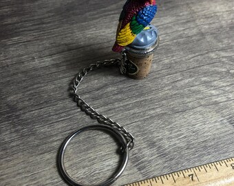 May include: A colourful parrot bottle stopper with a metal chain and ring. The parrot is perched on a cork stopper with a silver top.