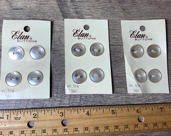 May include: Three cards of white plastic buttons with the text "Elan Buttons" and "HK, 514 50¢" printed on them. The buttons are silver and have four holes.