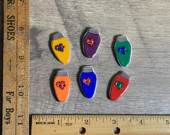 May include: Six ceramic lightbulb ornaments with a heart design in various colours, including red, orange, yellow, green, blue, and purple. The hearts are all slightly different and have a distressed look.
