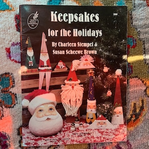 May include: A book titled "Keepsakes for the Holidays" by Charleen Stempel & Susan Scheewe Brown. The cover features illustrations of various holiday-themed crafts, including a Santa Claus face on a gourd, a snowman, and a gnome.