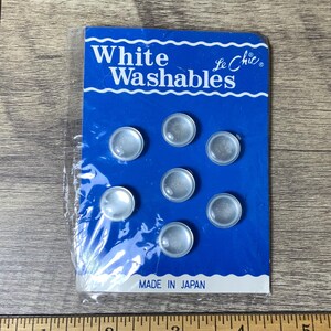 May include: A package of seven white, round, plastic buttons with a blue background card that reads "White Washables Le Chic" and "Made in Japan".