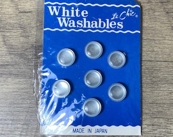 May include: A pack of seven white, round, plastic buttons with a blue background card that reads "White Washables Le Chic" and "Made in Japan".