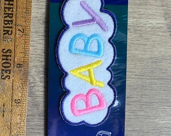 May include: A white felt patch with the word "BABY" embroidered in colourful letters. The letters are outlined in blue and the patch is packaged on a blue card with the words "Wrights Applique Application" and "Iron Ons Planchados Auto Fer Chaud".