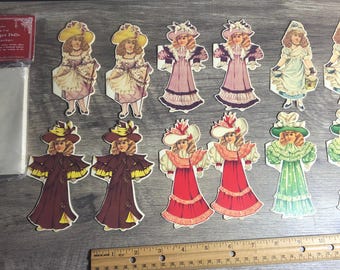 May include: A set of 10 paper dolls, featuring girls wearing various dresses and hats. The dolls are printed on cardstock and are ready to be cut out and played with. The dolls are all dressed in different colours and styles, including pink, blue, green, red, and yellow.