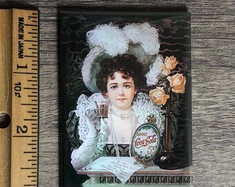 May include: A vintage Coca-Cola advert featuring a woman in a white hat and green dress holding a glass of Coca-Cola. The advert features the text "Drink Coca-Cola 5c".