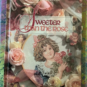 Pode incluir: Um livro intitulado "Sweeter Than the Rose" com um desenho de ponto cruz de uma menina segurando rosas. A capa inclui rosas e uma fita, com o texto "A Leisure Arts Publication".