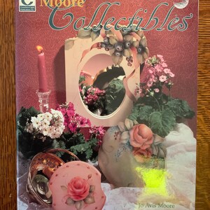 May include: A book cover titled "Moore Collectibles" featuring a decorative mirror, flowers, and a candle. The cover has a pink background with the author's name, Jo Avis Moore, at the bottom. The book is published by Golden Eagle Publications, Inc.