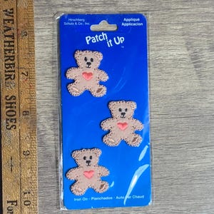 May include: A package of three iron-on patches featuring brown teddy bears with pink hearts. The package is blue and says "Patch it Up" in white letters.