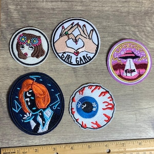 Pode incluir: Uma coleção de patches bordados com vários designs. Um apresenta um rosto com uma coroa de flores, outro diz "GIRL GANG", e um terceiro retrata um OVNI. Outros patches incluem um olho estilizado e um retrato.