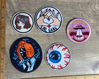May include: A selection of embroidered patches with diverse designs. One shows a face with a floral crown, another reads "GIRL GANG", and a third depicts a UFO. Other patches include a stylized eye and a portrait.