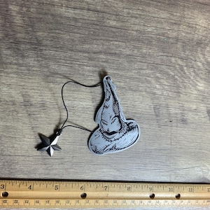 Può includere: Un ciondolo d'argento con un'illustrazione in bianco e nero del Cappello Parlante della serie Harry Potter. Il ciondolo è attaccato a un ciondolo a stella d'argento con uno stringa nera.