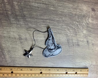 May include: A silver charm featuring a black and white illustration of the Sorting Hat from the Harry Potter series. The charm is attached to a silver star charm with a black string.