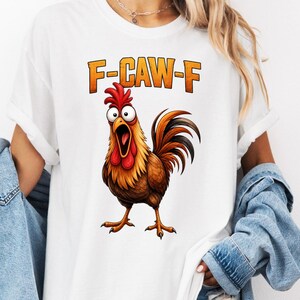 May include: A white t-shirt featuring a graphic of a brown rooster with wide, surprised eyes and an open beak. Above the rooster, the text F-CAW-F is printed in orange block letters. The shirt has short sleeves and a crew neckline.
