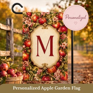 Könnte beinhalten: Eine rechteckige Gartenflagge mit einem cremefarbenen, verzierten Rahmen in der Mitte, der einen großen roten Buchstaben M enthält. Der Hintergrund ist mit einem Muster aus roten Äpfeln und rosa Apfelblüten verziert. Die Flagge hängt an einem schwarzen Metallständer.