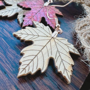 May include: Wooden leaf-shaped earrings with detailed vein patterns. The earrings are light brown with a darker brown outline and a gold-colored hook. One earring has a pink and purple gradient.
