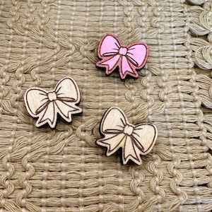 May include: Three wooden bow-shaped embellishments. Two are natural wood with dark brown outlines, and one is pink with glitter accents. The bows are arranged on a textured, beige surface.