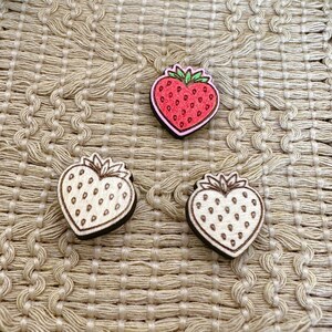 May include: Three wooden strawberry-shaped embellishments. One is red with a pink outline and green leaves, the other two are natural wood with brown outlines and small dot details. The embellishments are on a textured beige surface.