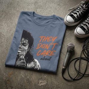 May include: Blue t-shirt with a black and white image of a singer holding a microphone, and the orange text "THEY DON'T CARE about". Black high-top trainers and a microphone with a cord are also in the image.