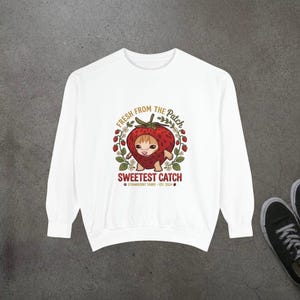 May include: A white long-sleeve sweatshirt featuring a graphic of a tabby cat inside a strawberry. The text reads FRESH FROM THE PATCH, SWEETEST CATCH, and STRAWBERRY TABBY - EST. 2024, surrounded by small strawberry vines and flowers.