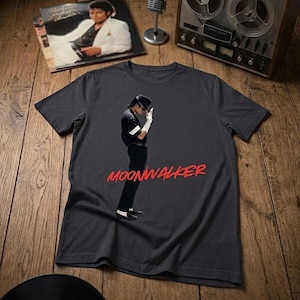 May include: Dark gray t-shirt featuring a graphic of a person in a black outfit and hat, with the word "MOONWALKER" in red. A vinyl record and album cover are visible in the background.