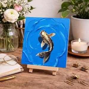 May include: A square painting of a koi fish in a blue water scene. The fish is gold and black, with detailed fins and a long tail. The painting is displayed on a small wooden easel, with a candle and flowers in the background.