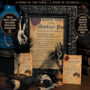 May include: A collection of three monkey paw props with black fur and twine wrapping, two mounted on wooden stands. Includes two printed relic cards titled Cursed Relic: The Monkey's Paw and a large framed display sign with the same text.