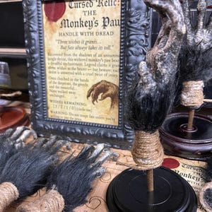Puede incluir: Una exhibición espeluznante con una pata de mono como accesorio, con un letrero enmarcado que dice "Cursed Relic: The Monkey's Paw". Las manos del accesorio son oscuras con pelo negro y bases envueltas en cuerda.