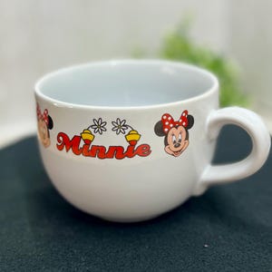 K&ouml;nnte beinhalten: Wei&szlig;e Minnie Mouse-Tasse aus Keramik mit abgerundeter Form und gro&szlig;em Henkel. Die Tasse zeigt das Wort "Minnie" in Rot sowie Bilder von Minnie Mouse und Blumenschmuck. F&uuml;r Getr&auml;nke konzipiert.