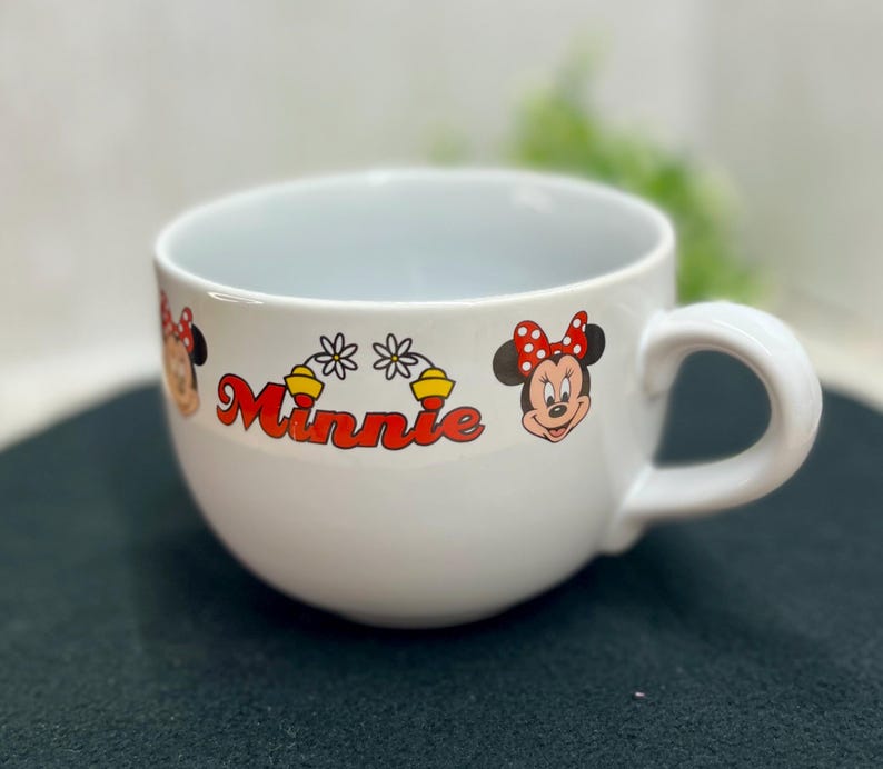 K&ouml;nnte beinhalten: Wei&szlig;e Minnie Mouse-Tasse aus Keramik mit abgerundeter Form und gro&szlig;em Henkel. Die Tasse zeigt das Wort "Minnie" in Rot sowie Bilder von Minnie Mouse und Blumenschmuck. F&uuml;r Getr&auml;nke konzipiert.