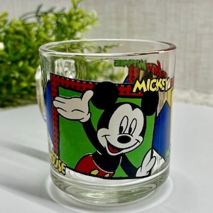 Peut inclure: Mug en verre transparent avec un dessin anim&eacute; de Mickey Mouse qui salue. La tasse a une poign&eacute;e et est d&eacute;cor&eacute;e du mot "Mickey" et d'autres &eacute;l&eacute;ments de dessins anim&eacute;s. L'arri&egrave;re-plan est flou, avec un feuillage vert visible.