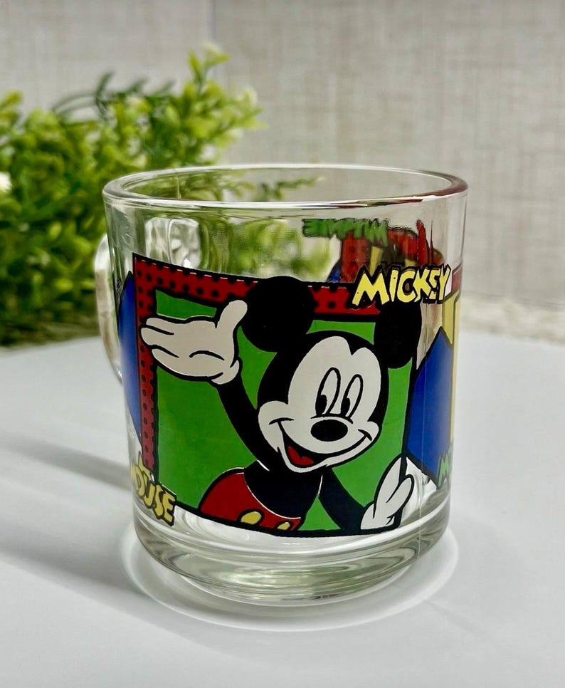 Peut inclure: Mug en verre transparent avec un dessin anim&eacute; de Mickey Mouse qui salue. La tasse a une poign&eacute;e et est d&eacute;cor&eacute;e du mot "Mickey" et d'autres &eacute;l&eacute;ments de dessins anim&eacute;s. L'arri&egrave;re-plan est flou, avec un feuillage vert visible.