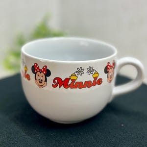 K&ouml;nnte beinhalten: Wei&szlig;er Keramikbecher mit Minnie Mouse-Motiven. Der Becher hat eine abgerundete Form mit Henkel und ist mit Minnie Mouse-Grafiken und dem Namen "Minnie" in roter Schrift verziert. Das Design beinhaltet Minnie's Gesicht und Schleife.