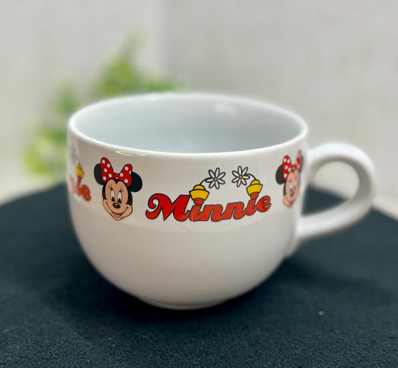 K&ouml;nnte beinhalten: Wei&szlig;er Keramikbecher mit Minnie Mouse-Motiven. Der Becher hat eine abgerundete Form mit Henkel und ist mit Minnie Mouse-Grafiken und dem Namen "Minnie" in roter Schrift verziert. Das Design beinhaltet Minnie's Gesicht und Schleife.