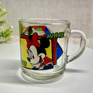 Peut inclure: Mug en verre transparent avec Minnie Mouse. Le design comprend le visage de Minnie, un n&oelig;ud rouge et le mot "MINNIE" en vert. La tasse a une anse et est adapt&eacute;e aux boissons.