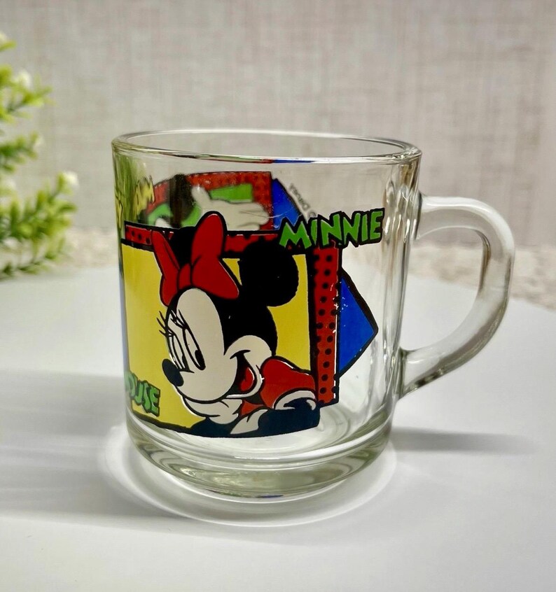 Peut inclure: Mug en verre transparent avec Minnie Mouse. Le design comprend le visage de Minnie, un n&oelig;ud rouge et le mot "MINNIE" en vert. La tasse a une anse et est adapt&eacute;e aux boissons.
