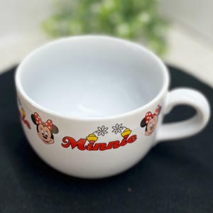 K&ouml;nnte beinhalten: Wei&szlig;er Keramikbecher mit Henkel, mit Minnie Mouse-Grafiken. Der Becher hat ein rot-gelbes "Minnie"-Textdesign sowie Minnie Mouses Gesicht und Schleife. Der Becher ist f&uuml;r Getr&auml;nke konzipiert.