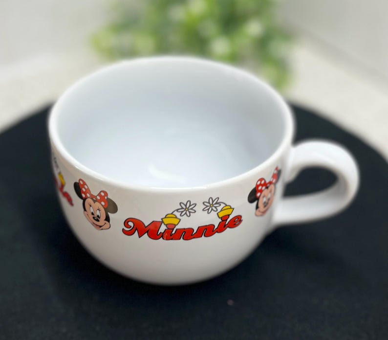 K&ouml;nnte beinhalten: Wei&szlig;er Keramikbecher mit Henkel, mit Minnie Mouse-Grafiken. Der Becher hat ein rot-gelbes "Minnie"-Textdesign sowie Minnie Mouses Gesicht und Schleife. Der Becher ist f&uuml;r Getr&auml;nke konzipiert.