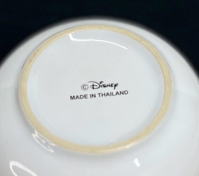 K&ouml;nnte beinhalten: Wei&szlig;er Keramiknapf mit hellem beigen Rand. Der Napf hat das Disney-Logo und den Text "MADE IN THAILAND" auf der Unterseite aufgedruckt.